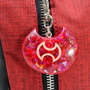 May include: A red, sparkly, resin charm with a white symbol in the shape of a crescent moon. The charm is attached to a silver zipper pull.