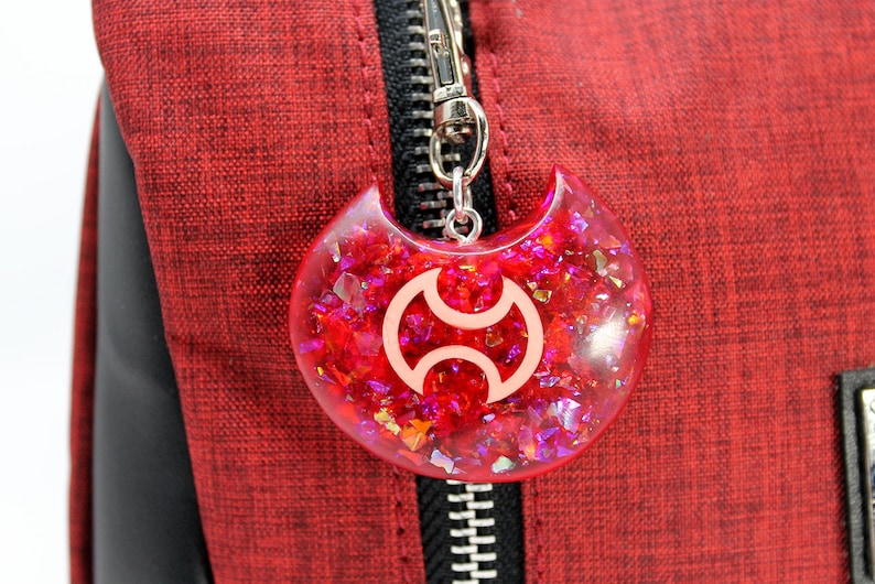 May include: A red, sparkly, resin charm with a white symbol in the shape of a crescent moon. The charm is attached to a silver zipper pull.