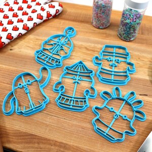 May include: Five blue plastic cookie cutters shaped like cartoon characters on a wooden surface. The cutters are in the shape of a ghost, a carousel, a snowman, a ghost with a flower on its head, and a ghost with a hat.
