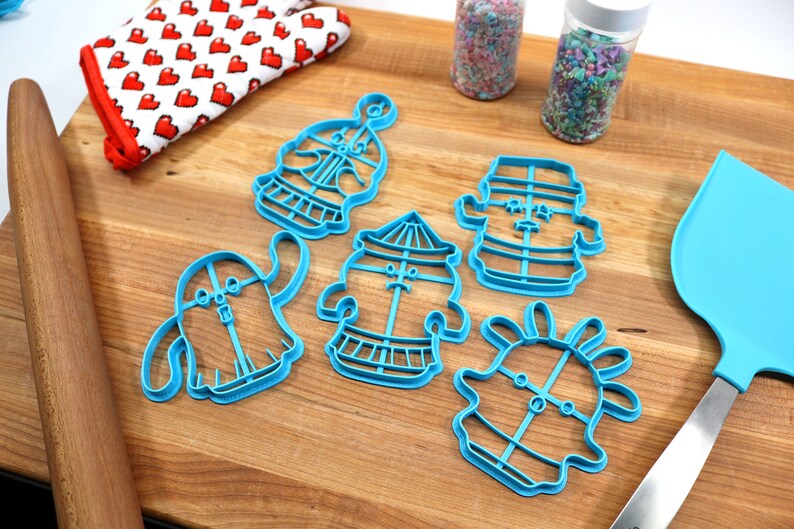 May include: Five blue plastic cookie cutters shaped like cartoon characters on a wooden surface. The cutters are in the shape of a ghost, a carousel, a snowman, a ghost with a flower on its head, and a ghost with a hat.