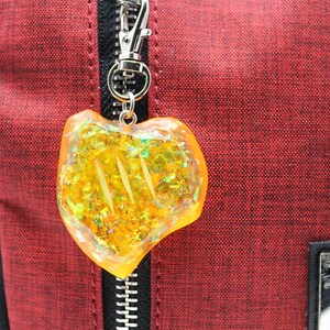 May include: A yellow heart-shaped charm with iridescent glitter embedded inside. The charm is attached to a silver zipper pull on a red bag.