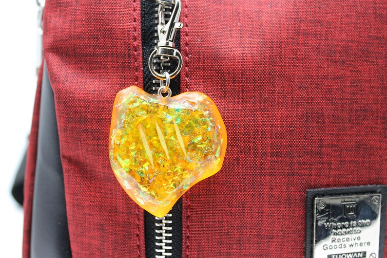May include: A yellow heart-shaped charm with iridescent glitter embedded inside. The charm is attached to a silver zipper pull on a red bag.