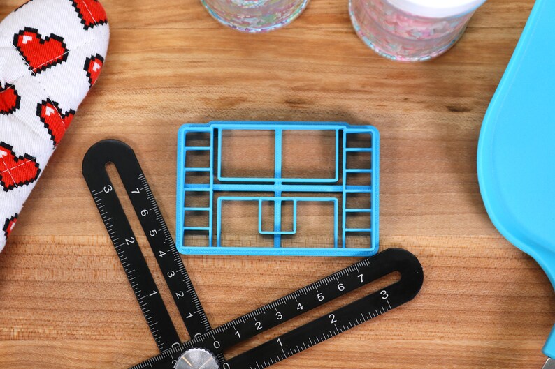 May include: A blue plastic cookie cutter shaped like a house with multiple windows. The cutter is on a wooden surface with a black ruler and a blue spatula.