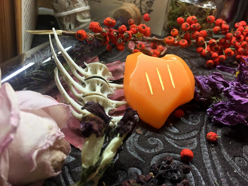 May include: Orange resin dice with three lines carved into the surface. The dice is surrounded by dried flowers, a glass vial with dried herbs, and a bone-like object.
