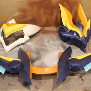 May include: A set of three blue, orange, and yellow plastic cosplay props. The props are shaped like a helmet, wings, and a sword.