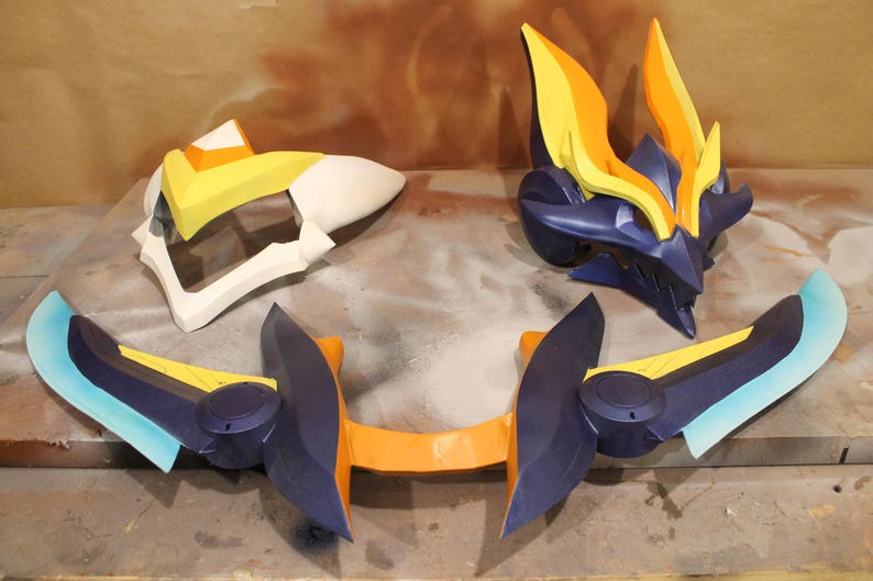 May include: A set of three blue, orange, and yellow plastic cosplay props. The props are shaped like a helmet, wings, and a sword.