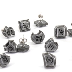 Puede incluir: Un conjunto de 12 pendientes plateados con varios s&iacute;mbolos y dise&ntilde;os. Los pendientes est&aacute;n hechos de metal y tienen un acabado texturizado.