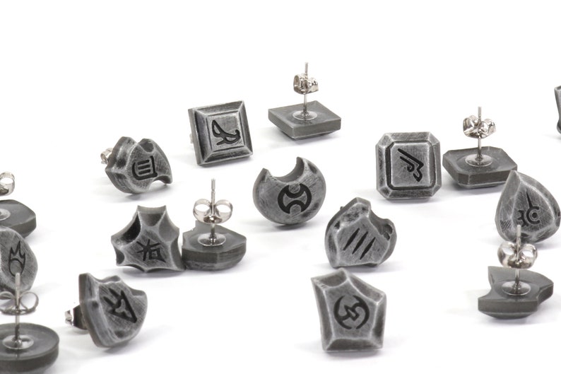 Puede incluir: Un conjunto de 12 pendientes plateados con varios s&iacute;mbolos y dise&ntilde;os. Los pendientes est&aacute;n hechos de metal y tienen un acabado texturizado.