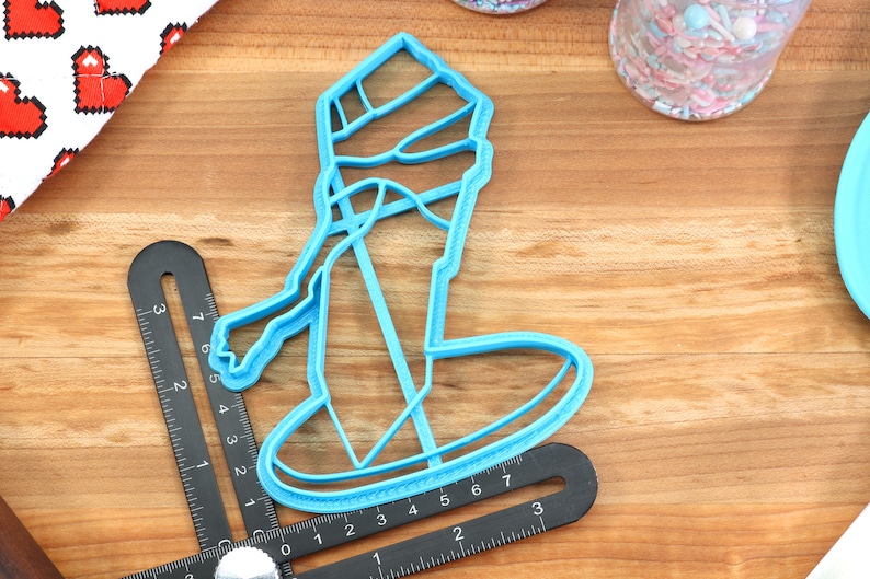 May include: A blue plastic cookie cutter shaped like a snowman wearing a hat and scarf. The cutter is on a wooden surface.