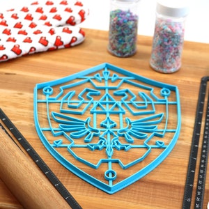 May include: Blue plastic cookie cutter shaped like the Triforce symbol from the Legend of Zelda video game series. The cutter is on a wooden surface with a rolling pin, measuring tape, and sprinkles.