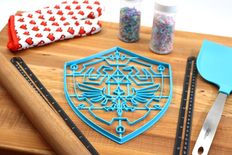 May include: Blue plastic cookie cutter shaped like the Triforce symbol from the Legend of Zelda video game series. The cutter is on a wooden surface with a rolling pin, measuring tape, and sprinkles.
