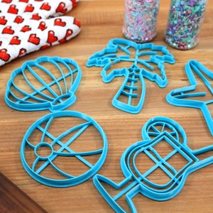 May include: A set of six blue plastic cookie cutters shaped like a seashell, palm tree, starfish, beach ball, cocktail glass, and a slice of lemon. The cutters are on a wooden surface.