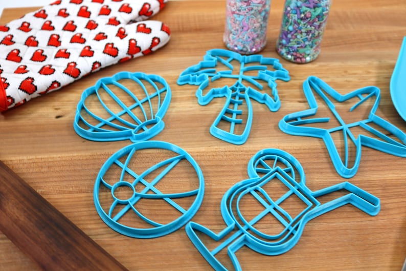 May include: A set of six blue plastic cookie cutters shaped like a seashell, palm tree, starfish, beach ball, cocktail glass, and a slice of lemon. The cutters are on a wooden surface.
