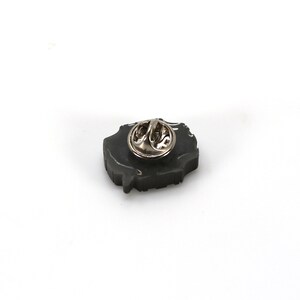May include: A black enamel pin with a detailed design. The pin has a silver metal clasp on the back.