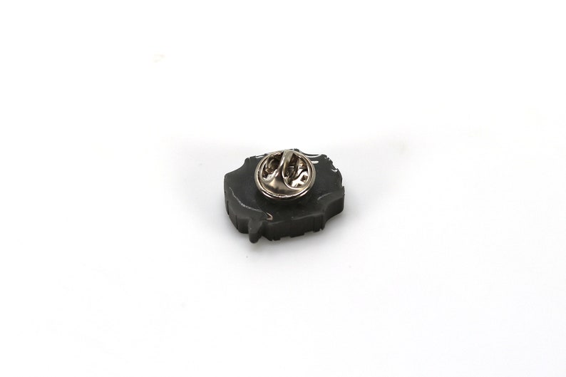 May include: A black enamel pin with a detailed design. The pin has a silver metal clasp on the back.