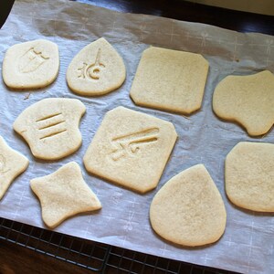 May include: A sheet of parchment paper with nine plain sugar cookies, each with a different design carved into the top. The cookies are arranged in a grid pattern.