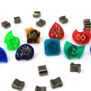 May include: A collection of colourful resin dice with various symbols and designs. The dice are arranged in a circle on a white surface, with several small metal pieces scattered around them.