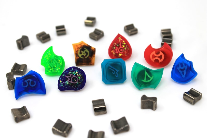 May include: A collection of colourful resin dice with various symbols and designs. The dice are arranged in a circle on a white surface, with several small metal pieces scattered around them.