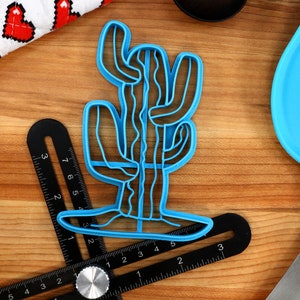 May include: A blue plastic cactus cookie cutter. The cutter is shaped like a tall cactus with multiple arms. The cutter is on a wooden surface.