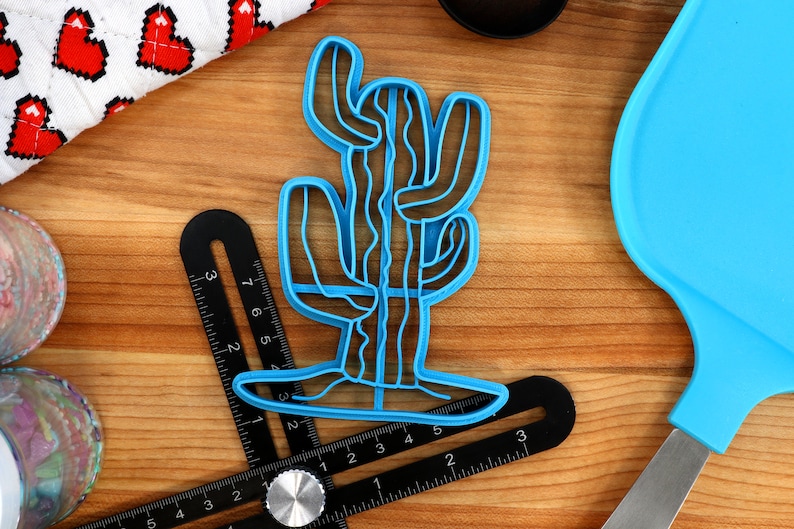 May include: A blue plastic cactus cookie cutter. The cutter is shaped like a tall cactus with multiple arms. The cutter is on a wooden surface.