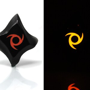 May include: A black, diamond-shaped, glow-in-the-dark dice with a red swirl design. The dice glows yellow in the dark.