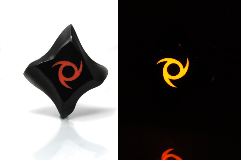 May include: A black, diamond-shaped, glow-in-the-dark dice with a red swirl design. The dice glows yellow in the dark.