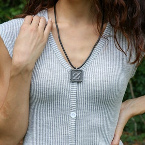 Pu&ograve; includere: Una collana d'argento con un ciondolo quadrato che presenta una lettera Z stilizzata. La collana &egrave; indossata da una persona con i capelli lunghi castani che indossa una camicia grigia a righe.
