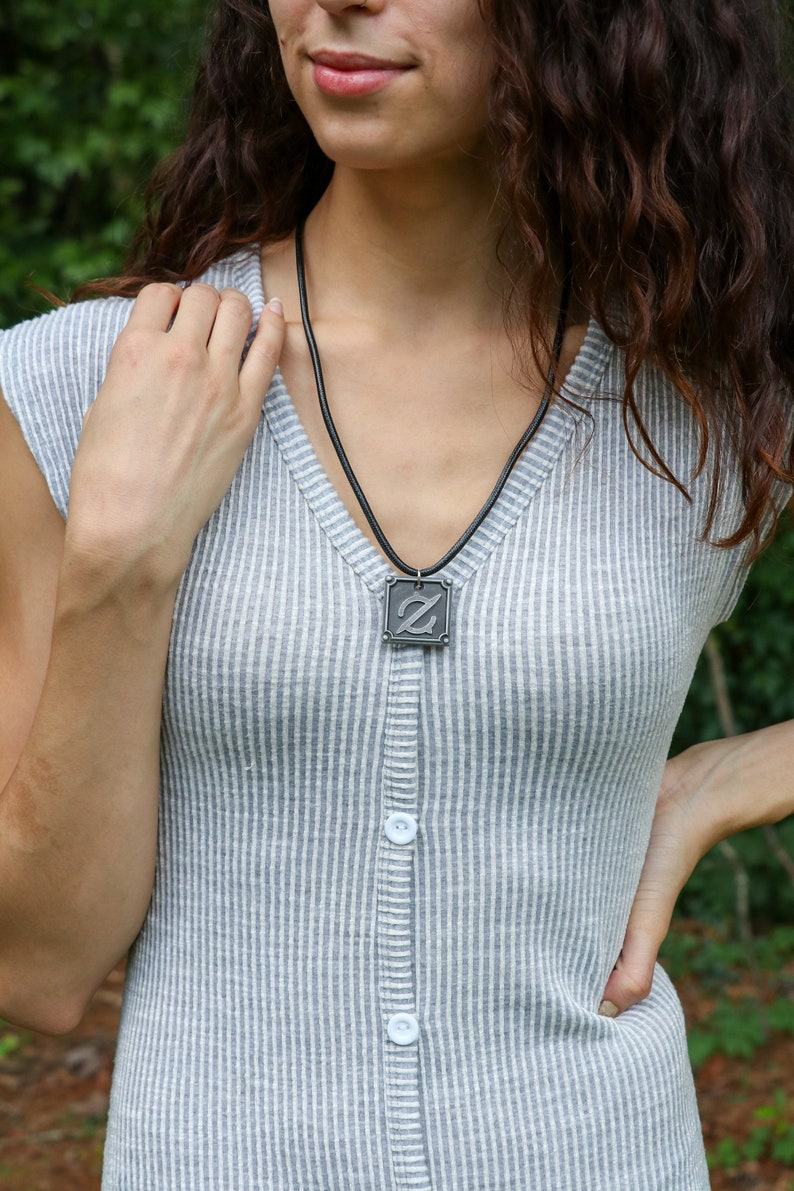 Pu&ograve; includere: Una collana d'argento con un ciondolo quadrato che presenta una lettera Z stilizzata. La collana &egrave; indossata da una persona con i capelli lunghi castani che indossa una camicia grigia a righe.