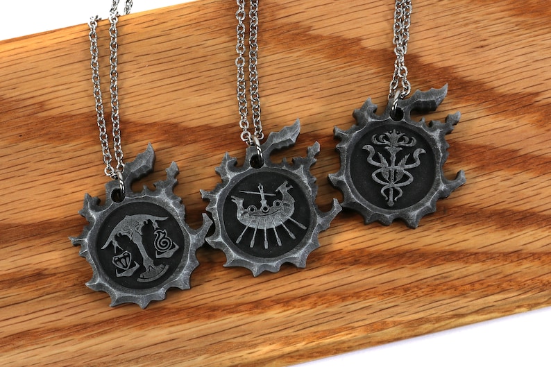May include: Three silver necklaces with pendants featuring intricate designs. The pendants are circular and have a textured, aged look. Each pendant features a different design, including a ship, a scale, and a stylized flower.