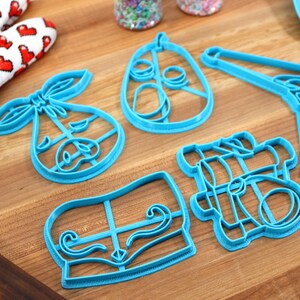 May include: A collection of five turquoise plastic cookie cutters in various shapes, including a baby carriage, a pear, and a mustache. A red and white heart-patterned oven mitt is visible in the background, suggesting a baking theme.