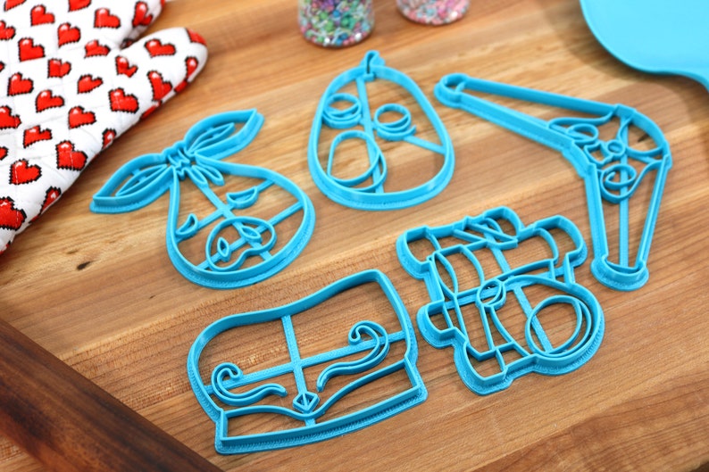 May include: A collection of five turquoise plastic cookie cutters in various shapes, including a baby carriage, a pear, and a mustache. A red and white heart-patterned oven mitt is visible in the background, suggesting a baking theme.