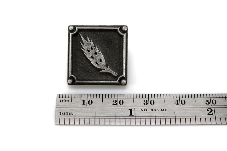 May include: A silver-toned metal square pin with a wheat stalk design. The pin is about 1 inch wide and is shown next to a ruler.
