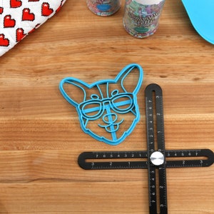 Pu&ograve; includere: Tagliapasta in plastica blu a forma di cane con occhiali da sole. Il tagliapasta &egrave; su una superficie di legno con un righello nero.