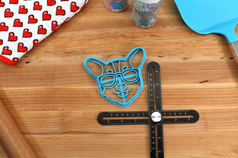 Pu&ograve; includere: Tagliapasta in plastica blu a forma di cane con occhiali da sole. Il tagliapasta &egrave; su una superficie di legno con un righello nero.