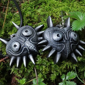 May include: Two black and silver Majora's Mask pendants, one on a black cord and the other on a silver chain. The masks are detailed with wood grain and spikes.
