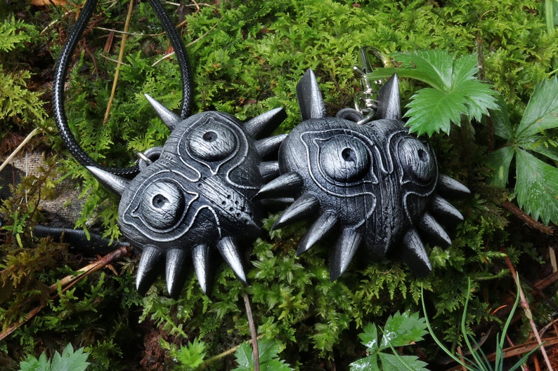 May include: Two black and silver Majora's Mask pendants, one on a black cord and the other on a silver chain. The masks are detailed with wood grain and spikes.