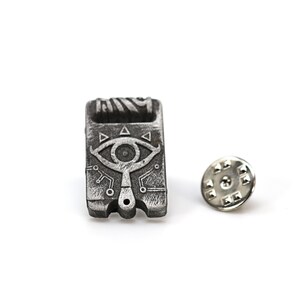 May include: A silver metal pin with a detailed design of a stylized eye with a triangular pattern. The pin is attached to a silver metal backing.