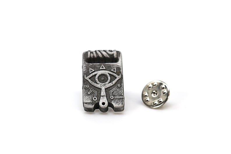 May include: A silver metal pin with a detailed design of a stylized eye with a triangular pattern. The pin is attached to a silver metal backing.