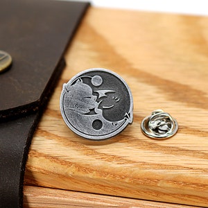 May include: A silver yin yang pin with a black cat and a white bat design. The pin is on a wooden surface with a brown leather strap.