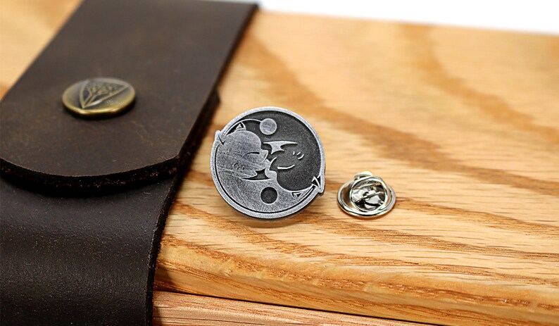 May include: A silver yin yang pin with a black cat and a white bat design. The pin is on a wooden surface with a brown leather strap.