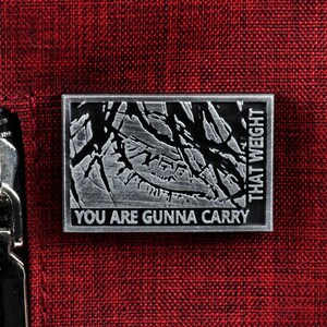 May include: A silver metal pin with a textured design and the text "You are gunna carry that weight".