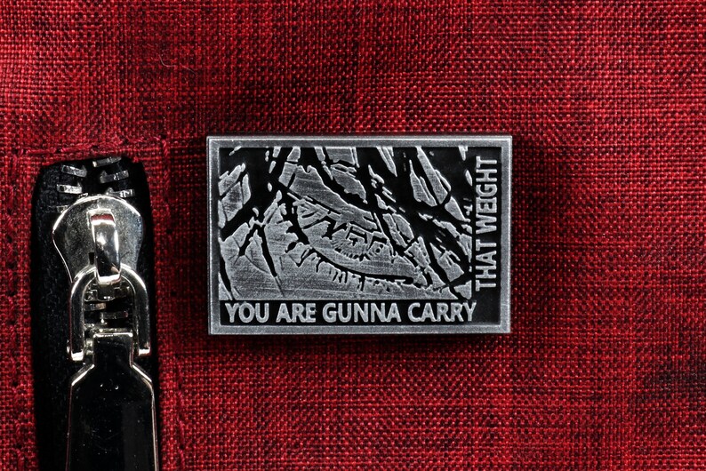 May include: A silver metal pin with a textured design and the text "You are gunna carry that weight".
