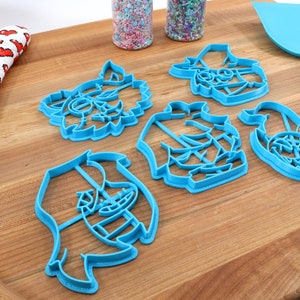 May include: Five blue plastic cookie cutters shaped like cartoon characters on a wooden surface. The cutters are in the shape of a werewolf, a pirate, a ghost, a pumpkin, and a witch.
