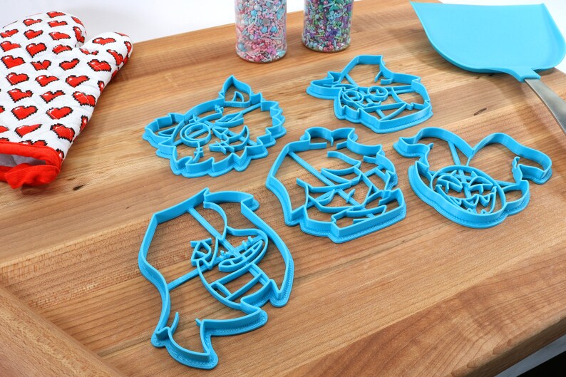 May include: Five blue plastic cookie cutters shaped like cartoon characters on a wooden surface. The cutters are in the shape of a werewolf, a pirate, a ghost, a pumpkin, and a witch.