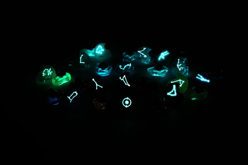 May include: A collection of glowing dice with constellations printed on each face. The dice are a variety of colors, including green, blue, and purple. The constellations are visible in the dark.