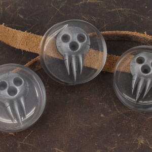 May include: Three clear, round resin cabochons with a gray skull design. The skulls have three eye holes and three pointed teeth. A tan leather strap is visible in the background, set against a brown leather surface.