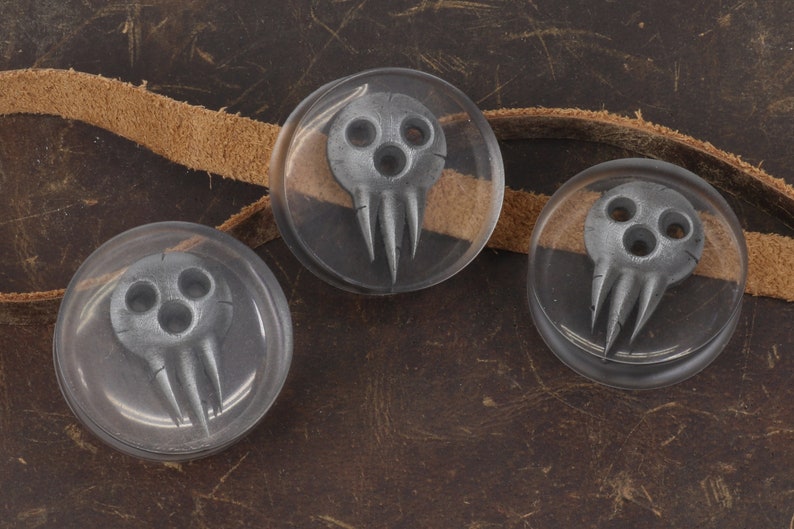 May include: Three clear, round resin cabochons with a gray skull design. The skulls have three eye holes and three pointed teeth. A tan leather strap is visible in the background, set against a brown leather surface.