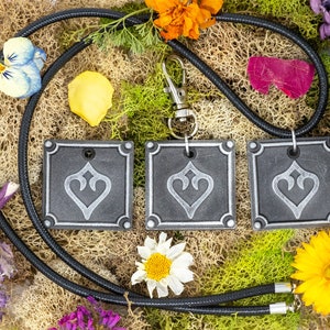 May include: Three gray square pendants with a silver heart design hang from a black cord with a silver clasp. The pendants are surrounded by dried flowers and moss.