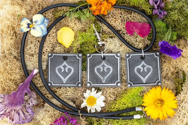 May include: Three gray square pendants with a silver heart design hang from a black cord with a silver clasp. The pendants are surrounded by dried flowers and moss.