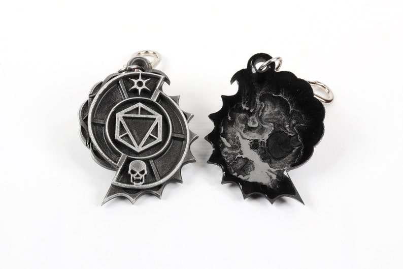 May include: Two silver metal keychains. One keychain features a circular design with a skull, a die, and a star. The other keychain is black and features a skull design.